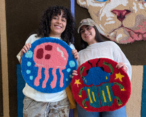 Two people holding hand tufted rugs one is a bug and the other is a jelly fish